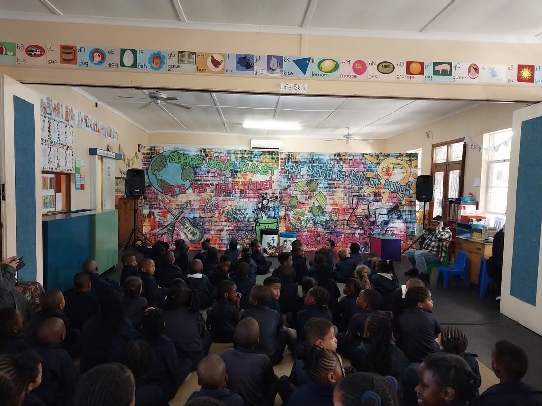 A group of young students in dark uniforms sit on the floor of a classroom facing a colorful mural, with a teacher at the front near a small table and learning materials—an inspiring scene from Bellville Preparatory School photos.