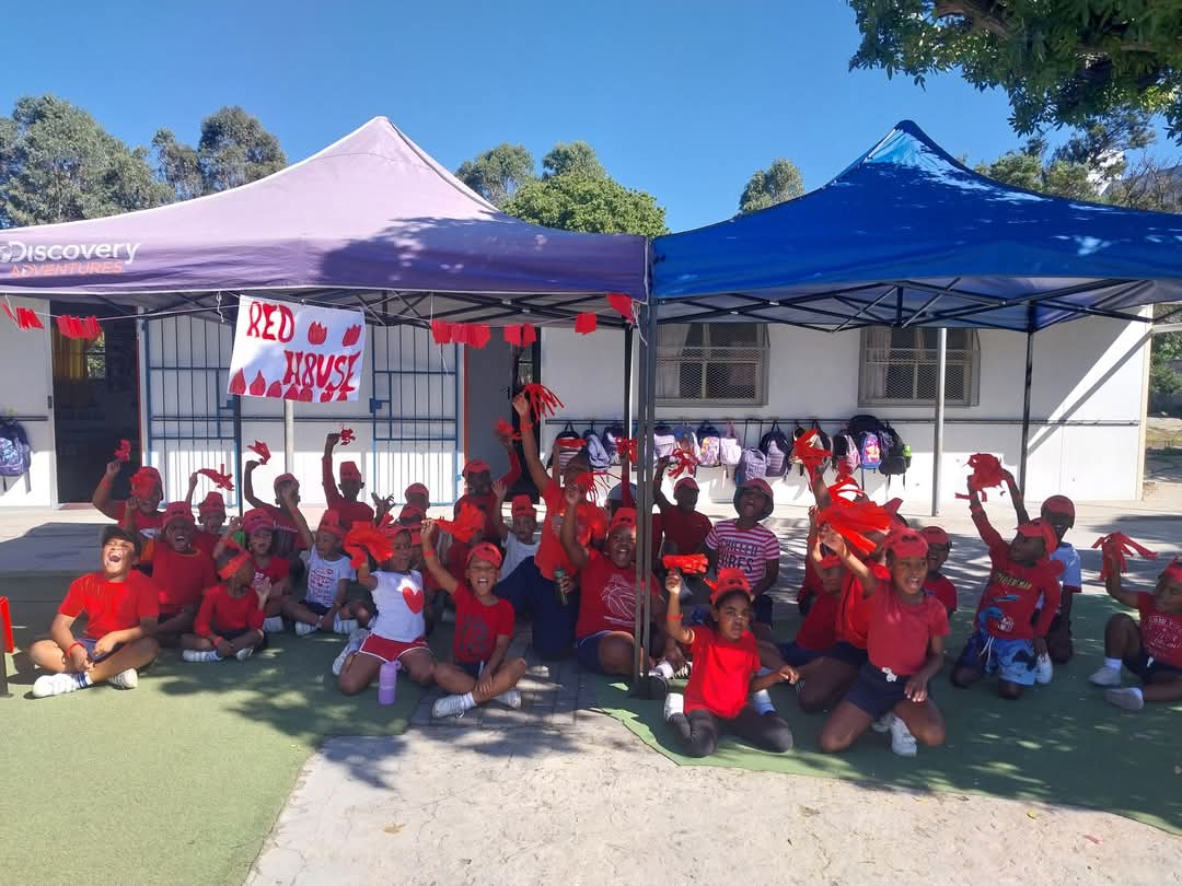 A group of children and adults in red clothes sit and stand under two canopies outdoors, holding up red items. A "Red House" sign hangs between the canopies. Trees and a building appear in the background of these Bellville Preparatory School photos.