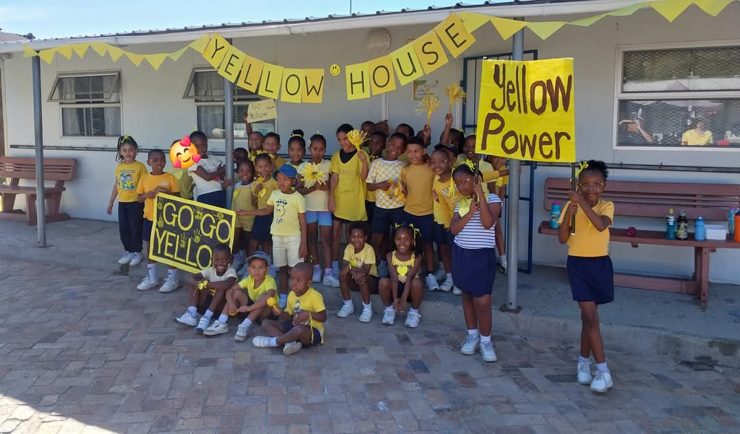 A group of children in yellow and blue outfits pose outside under a “Yellow House” banner in this Bellville Preparatory School photo. Some hold yellow signs reading “Yellow Power” and “Go Go Yellow,” with benches and a building in the background.