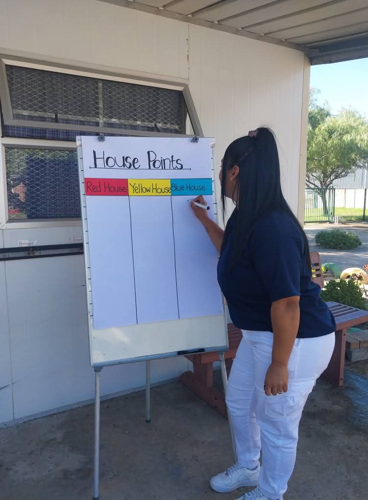 A person stands in front of a whiteboard with columns labeled Red House, Yellow House, and Blue House, writing on the board under an awning outside a building at Bellville Preparatory School.