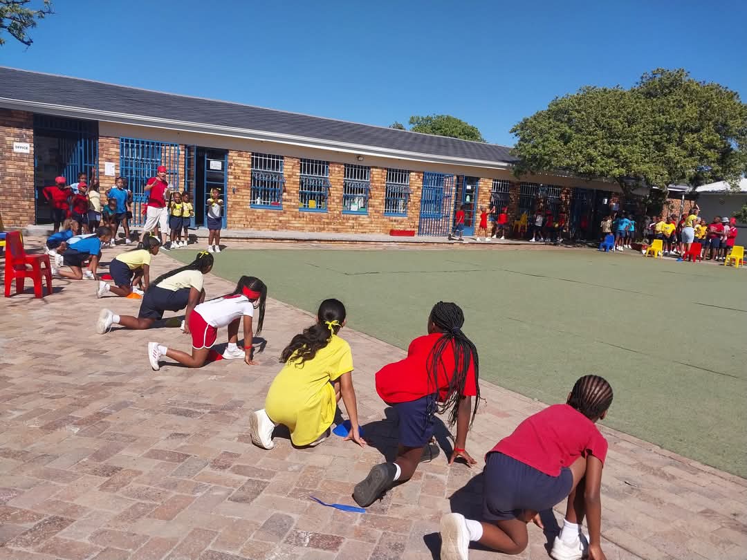 A group of children in sports uniforms crouch at a starting line on a paved area, preparing for a race, while more people gather near the brick school building—capturing the vibrant spirit found in Bellville Preparatory School photos.