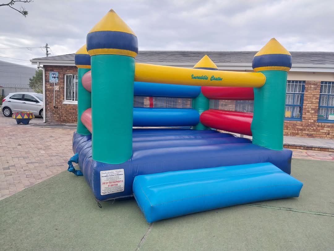 A colorful inflatable bounce house with blue, green, yellow, and red sections stands outdoors on a green mat near a brick building with barred windows—perfect for lively Bellville Preparatory School photos.