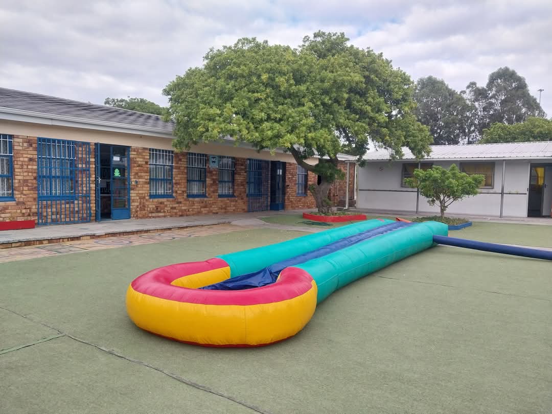 A colorful inflatable slip-and-slide is set up on artificial grass outside a brick school building at Bellville Preparatory School, with blue window bars and doors. Trees and another white building appear in the background under a cloudy sky.