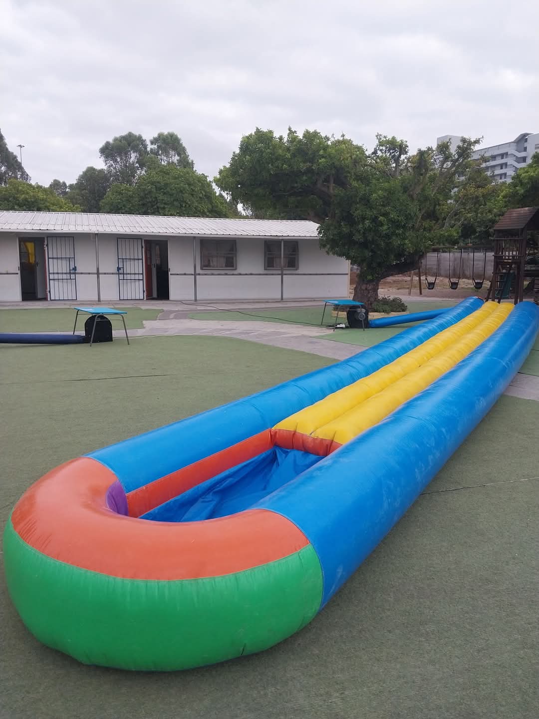 An inflatable slip-and-slide ride with red, green, blue, and yellow sections is set up on a grassy area near a building at Bellville Preparatory School, with trees and a playground visible in the background.