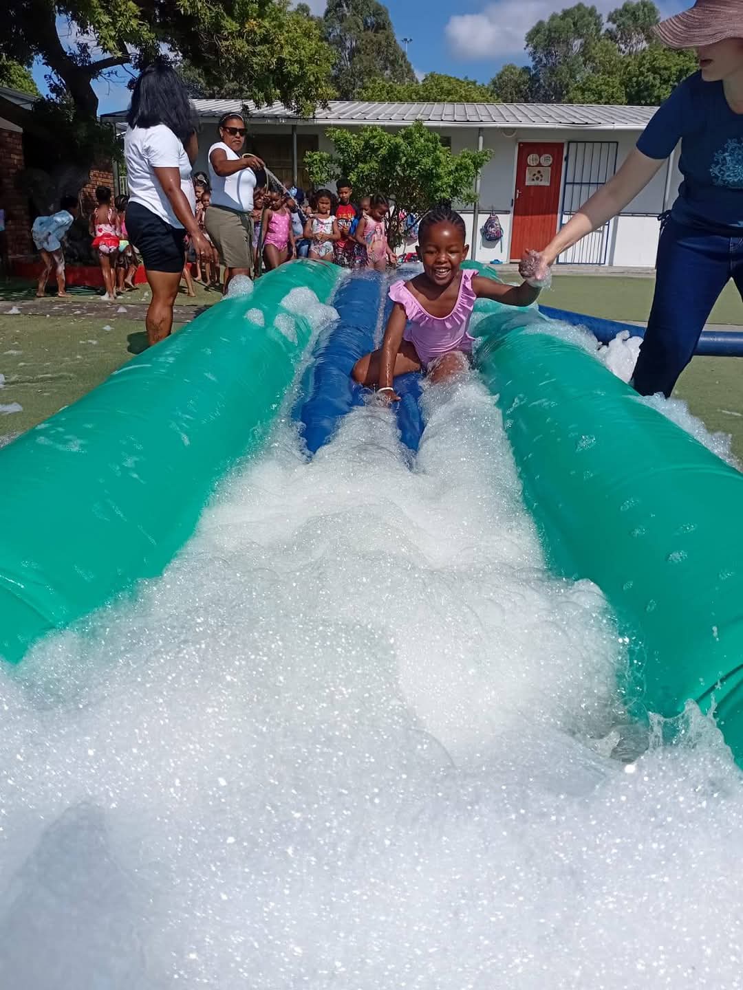 A young girl slides down a foam-covered inflatable slip-and-slide outdoors while an adult assists her; other children and adults are visible in the background on a sunny day at Bellville Preparatory School, as captured in these photos.