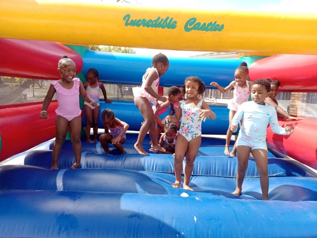 A group of young children wearing swimsuits play and jump on a large blue and red inflatable bouncy castle with the words “Incredible Castles” written at the top, enjoying a fun moment captured in Bellville Preparatory School photos.