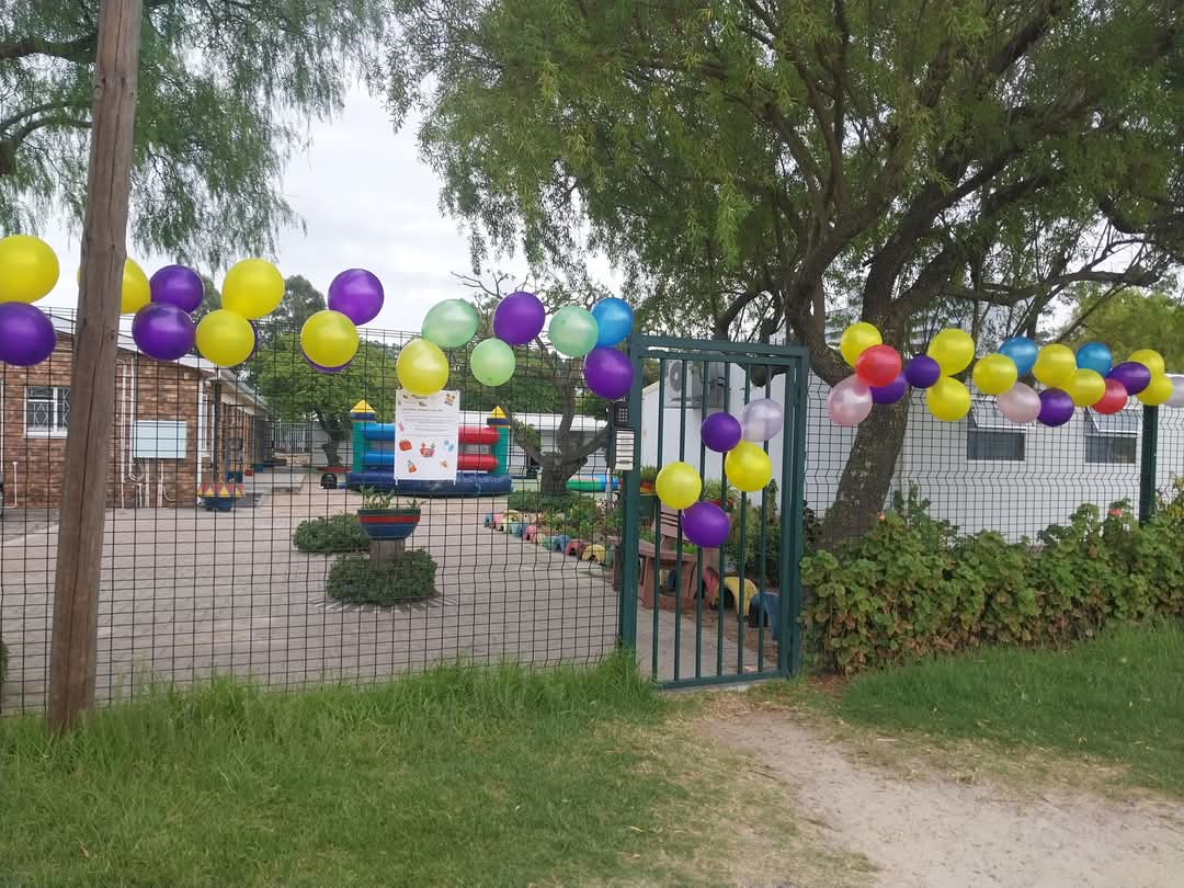 A metal gate and fence decorated with colorful balloons in yellow, purple, green, blue, and pink. Inside the fence are playground structures and potted plants on a concrete surface—a scene seen in many Bellville Preparatory School photos.
