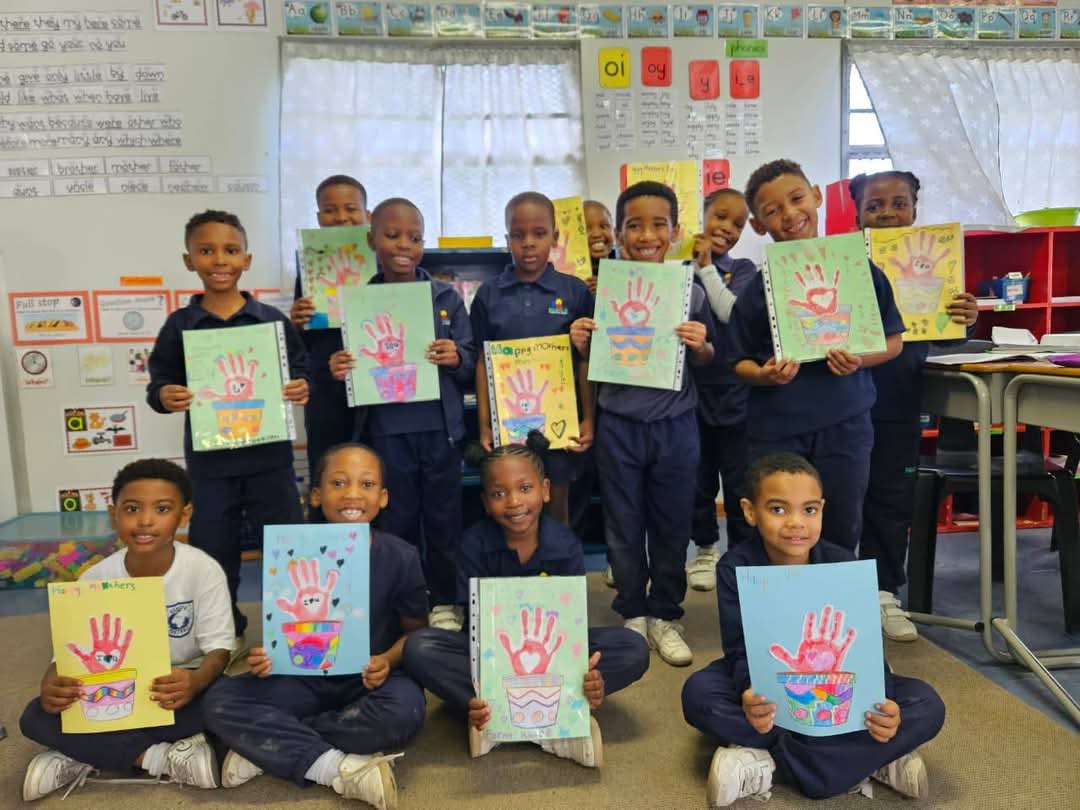 A group of schoolchildren in uniforms stand and kneel in a classroom, each holding up colorful handprint artwork. Bellville Preparatory School photos show walls decorated with charts, drawings, and educational posters.