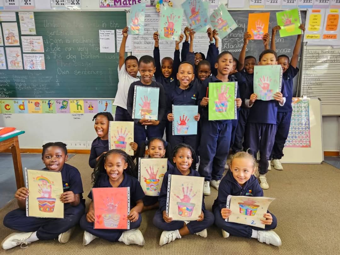 A group of school children in uniform pose in a classroom, smiling and holding up colorful handprint art projects. Behind them, chalkboards display writing and classroom posters are visible on the walls in these Bellville Preparatory School photos.