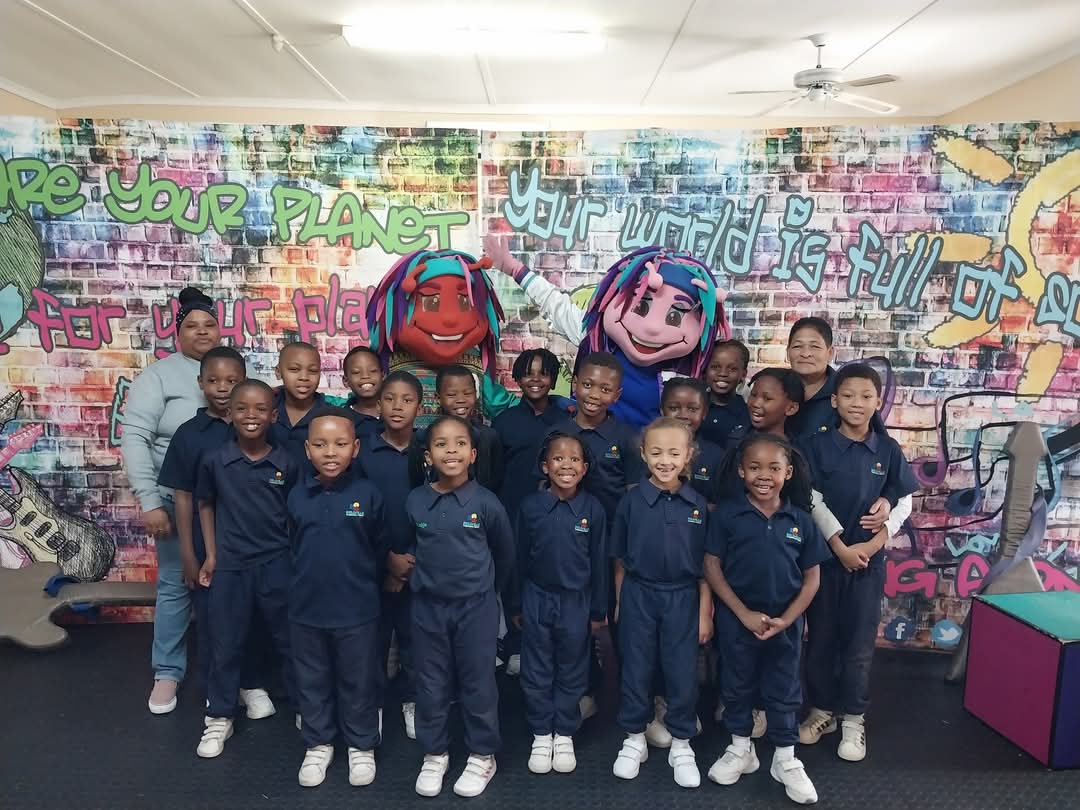 A group of school children in navy uniforms and two adults pose for a Bellville Preparatory School photo indoors, with two people in colorful character costumes and a graffiti-style mural in the background.