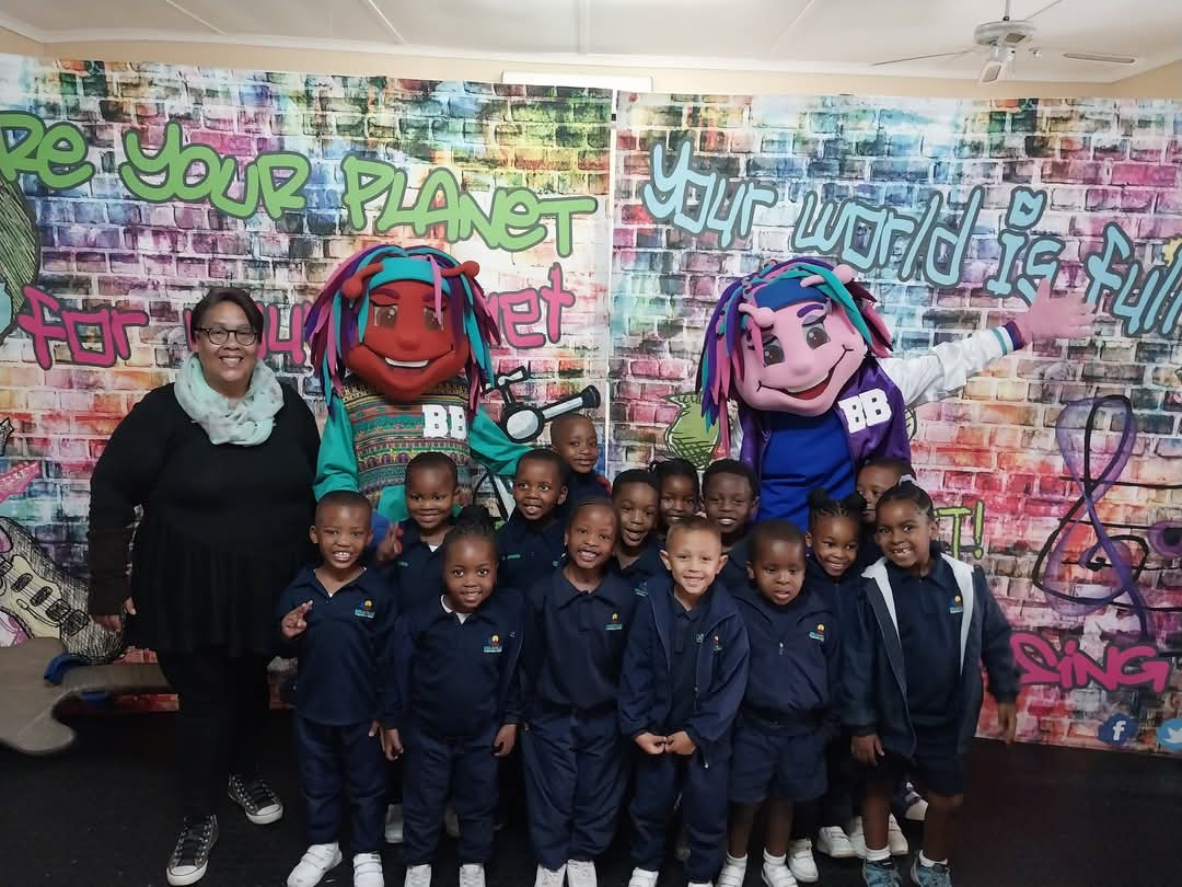 A group of young children in navy blue uniforms from Bellville Preparatory School stands in front of a colorful graffiti wall with two people in large character costumes and one adult woman on the left. All are posing and smiling for the photo.
