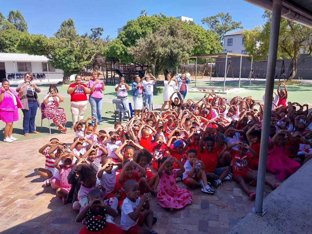 A large group of young children, dressed mostly in red, white, and pink, sit together outside on a paved area, making heart shapes with their hands—captured in one of the cheerful Bellville Preparatory School photos—while several adults join in behind them.
