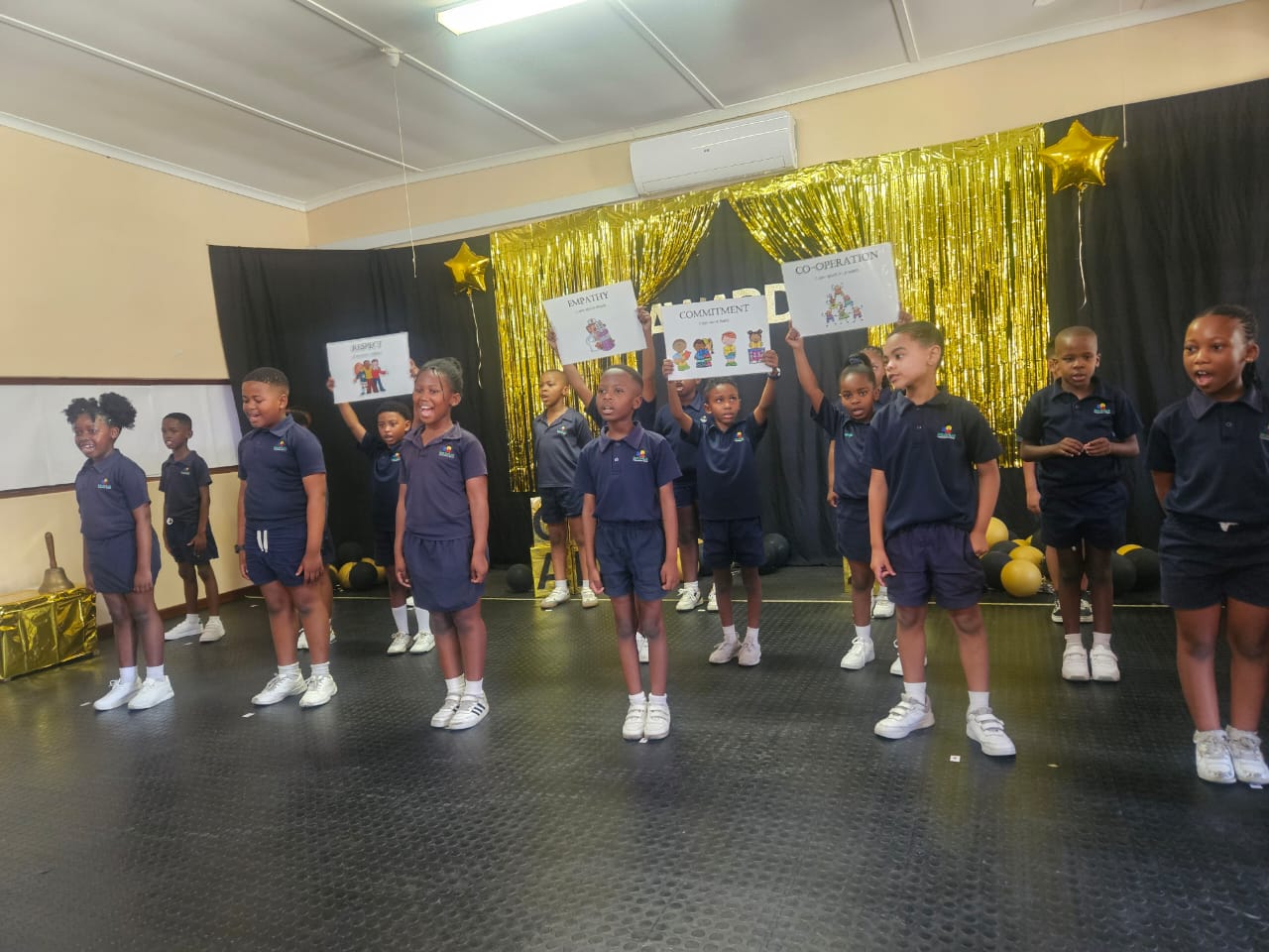 A group of young children in navy blue uniforms stand in rows on a stage decorated with gold stars, black and gold drapes, and posters displaying words like "Commitment" and "Cooperation.