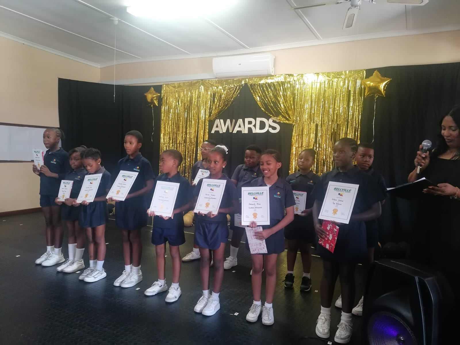 A group of young children in navy blue uniforms stand in a row on stage holding certificates. A woman stands to the right with a microphone. The backdrop says “AWARDS” with gold decorations and star-shaped balloons.