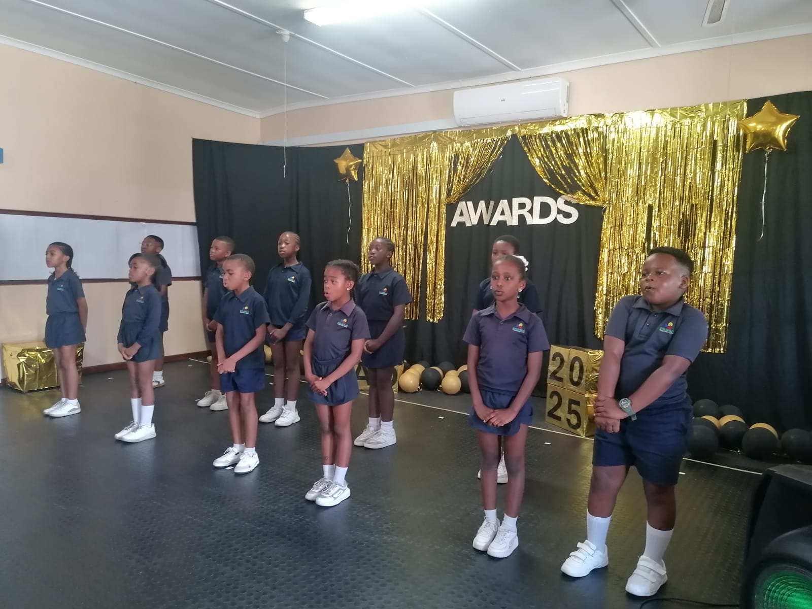 A group of children in matching uniforms stands in rows on a stage decorated with gold streamers, balloons, and a sign that reads "AWARDS." The children stand with hands in front, looking forward.