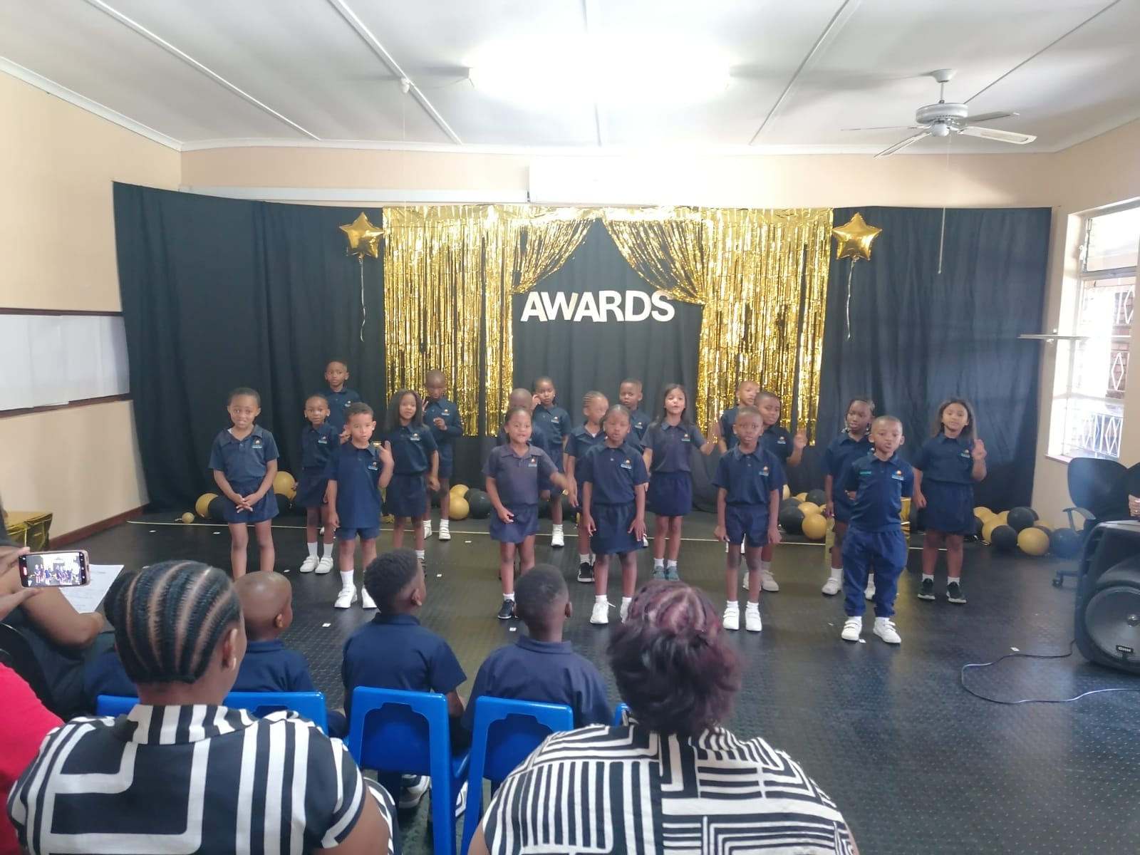 A group of children in navy uniforms stand on a stage with gold decorations and a black backdrop that says "AWARDS." Several adults are seated in the audience, watching the performance.