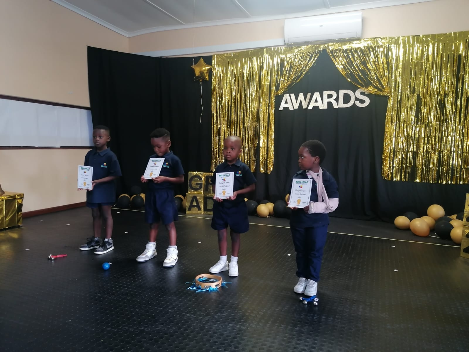 Four young children in school uniforms stand in a row on a stage holding certificates. The stage is decorated with gold streamers, balloons, and a sign that says "AWARDS" in large white letters.