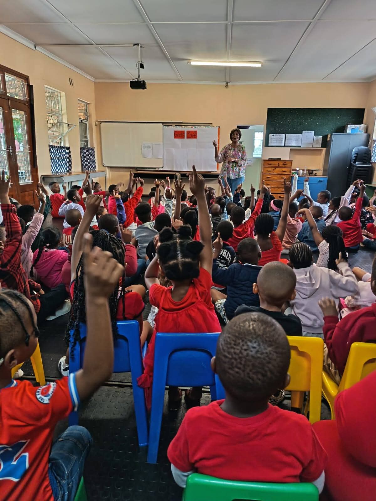 A classroom filled with young children seated on colorful chairs, raising their hands while a teacher stands at the front near a whiteboard and bulletin board. The students face forward, and sunlight shines through the windows.