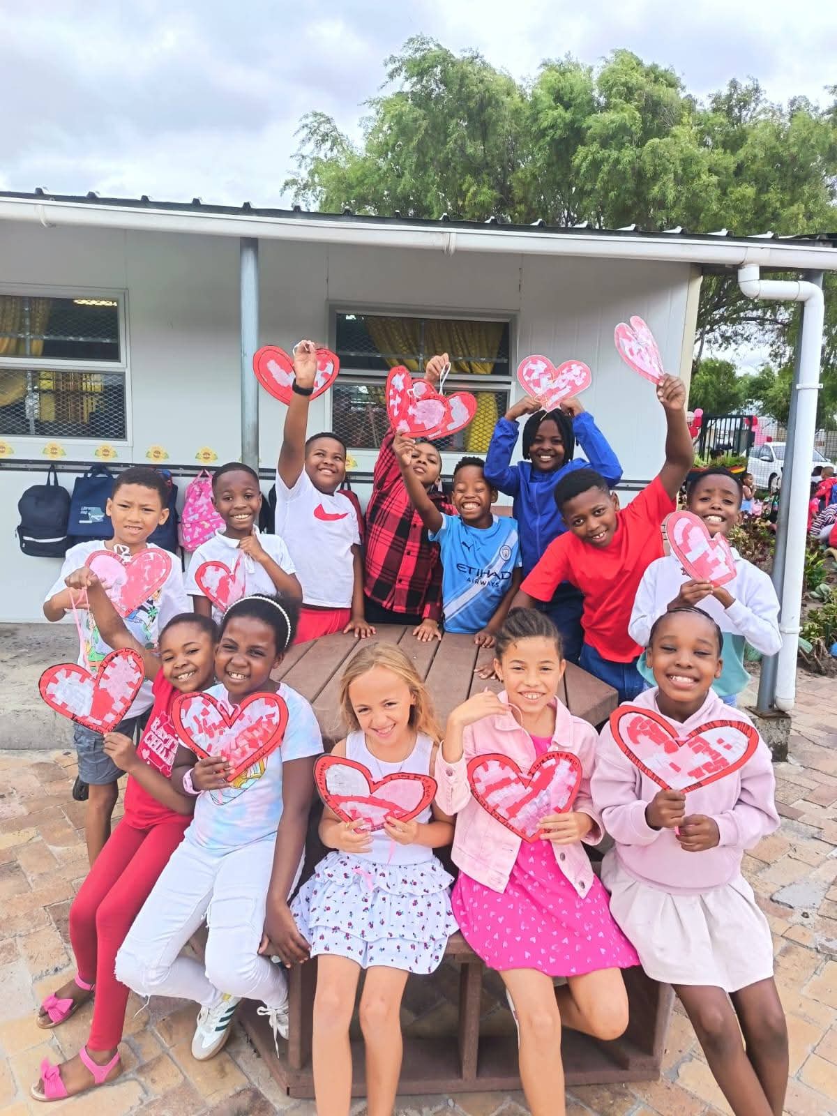 A group of children sit and stand around an outdoor table, smiling and holding decorated paper hearts. A building with windows and yellow curtains is in the background, and trees and plants surround the area.