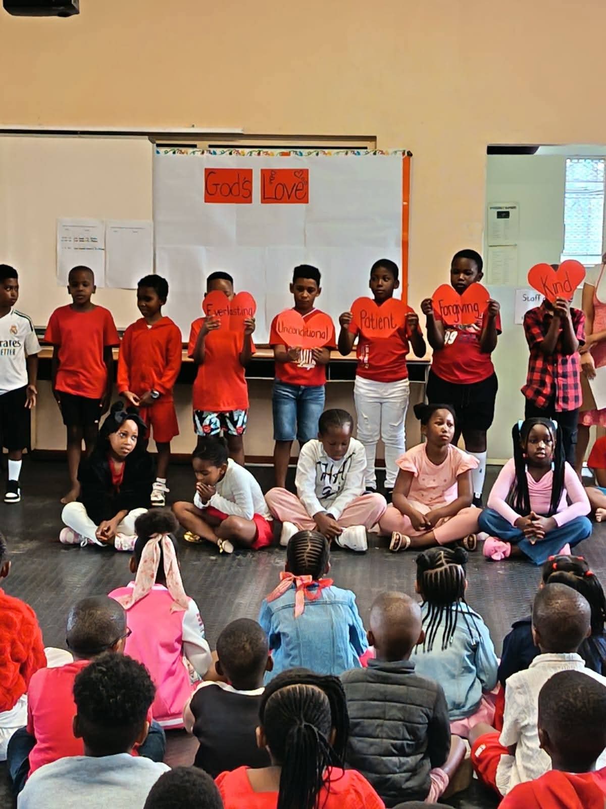 A group of children are standing and sitting in a classroom. The standing children hold large red heart-shaped signs with words like "Everlasting," "Unconditional," "Patient," "Forgiving," and "Kind.