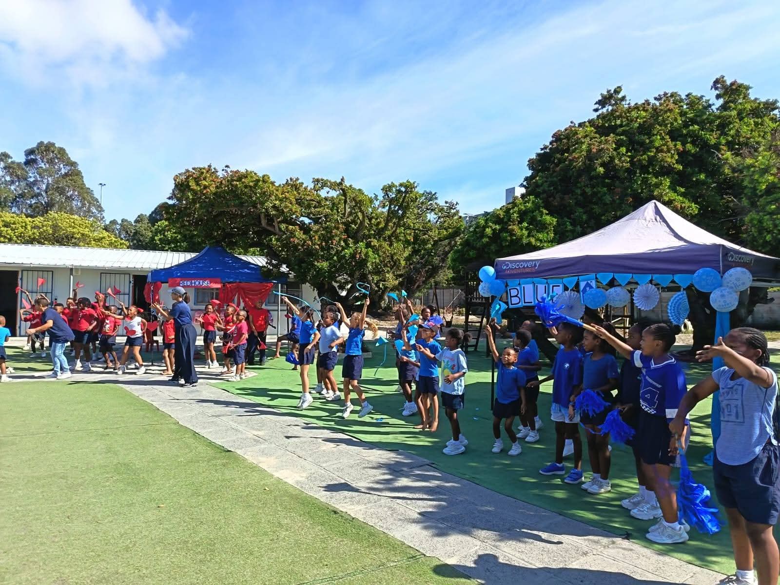 Children in red and blue shirts stand in groups on a sports field, with blue and red tents in the background. Blue decorations hang from one tent. Adults are present, and trees line the area under a blue sky.