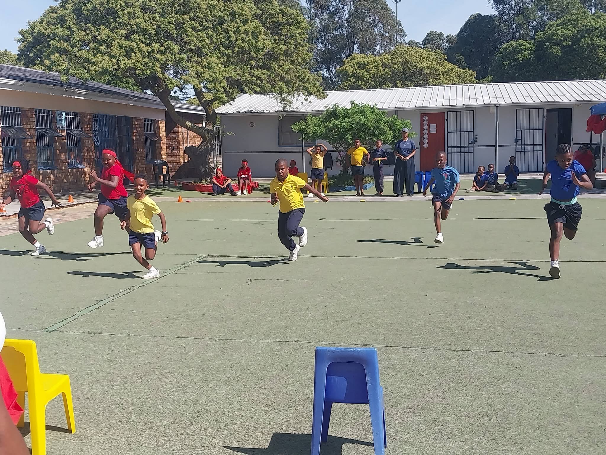 A group of children in sports uniforms are running a race on a paved outdoor area, with trees, buildings, and other people visible in the background. Colored plastic chairs are placed along the race track.