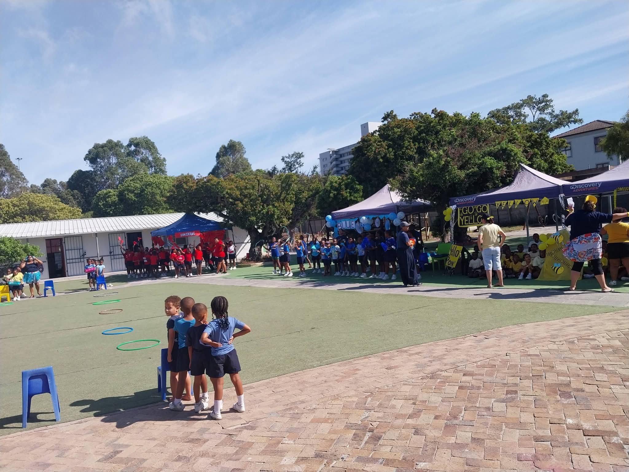 Children in different colored uniforms stand in groups near tents on a school playground. Some children are gathered near hoops on the ground, while adults and more children are seated or standing by the tents under a blue sky.