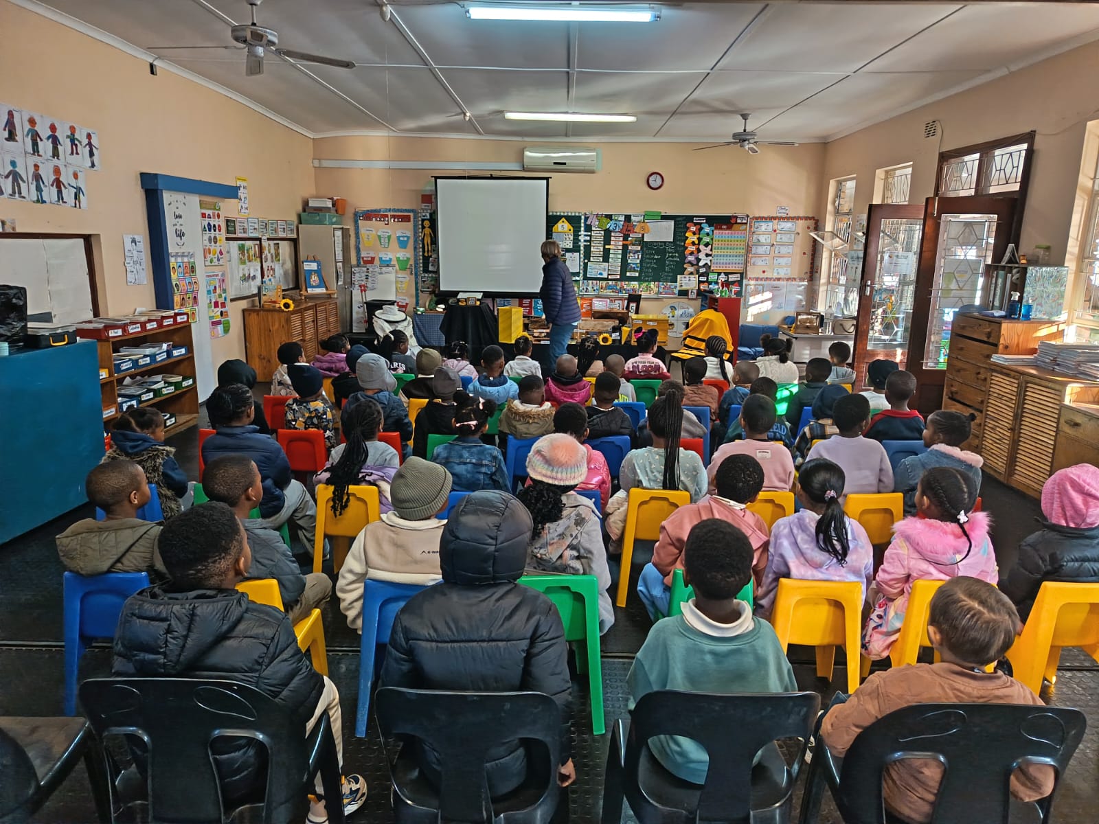 A group of children sit on colorful chairs facing a teacher at the front of a classroom. The room has educational posters, shelves, and a projector screen. The children are dressed warmly and attentive.