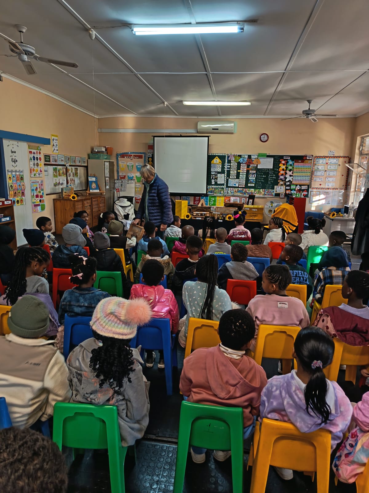 A classroom filled with young children sitting on colorful chairs facing a teacher at the front, with educational posters and a whiteboard visible on the walls.