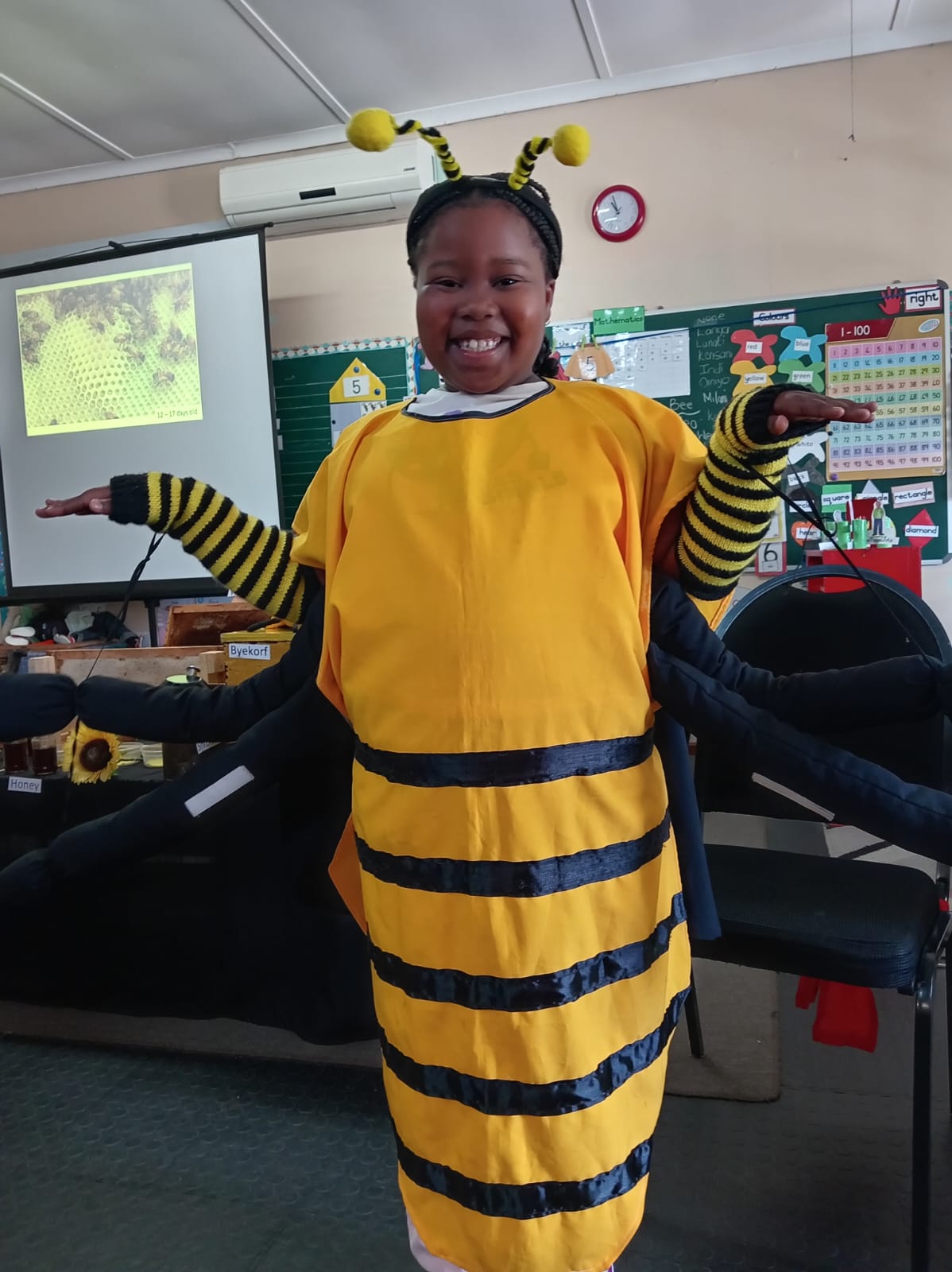 A child in a yellow and black bee costume stands in a classroom, smiling with arms raised. The classroom has colorful posters on the wall, a clock, and a projector displaying a honeycomb image.