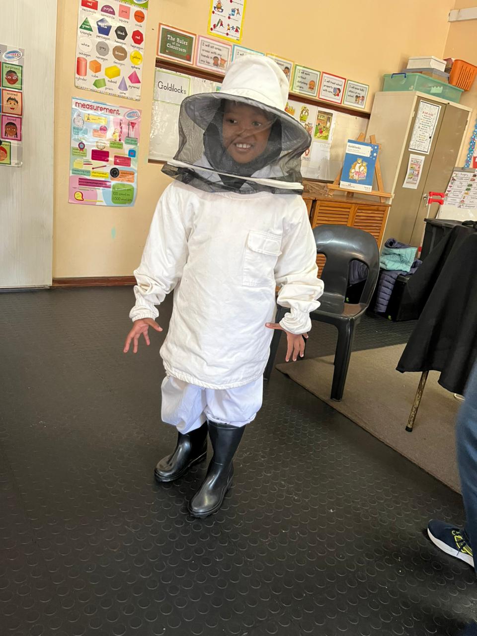 A child is standing indoors wearing a white beekeeping suit, black boots, and a protective hat with a mesh veil. Classroom posters and educational materials are visible on the walls in the background.