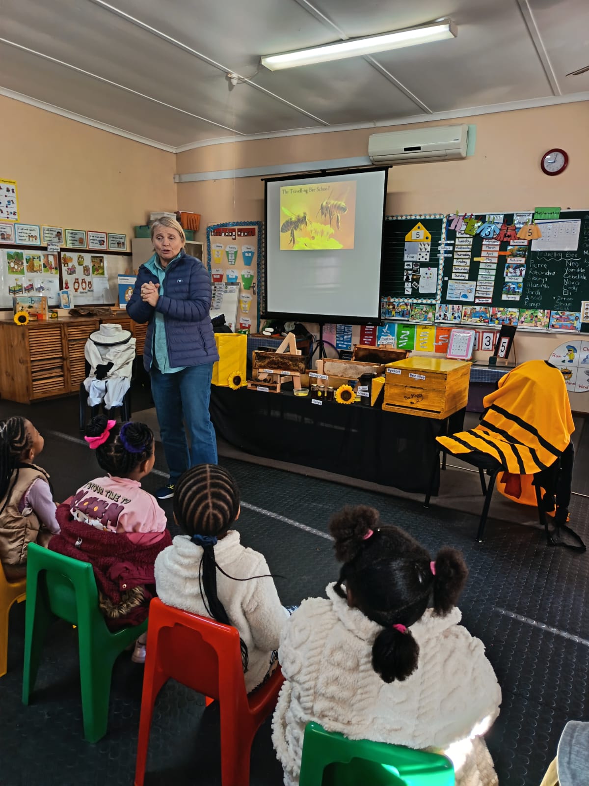 A woman presents to a group of young children in a classroom, showing an object in her hand. Behind her are beekeeping materials, sunflowers, and a projector screen displaying a bee-themed presentation.