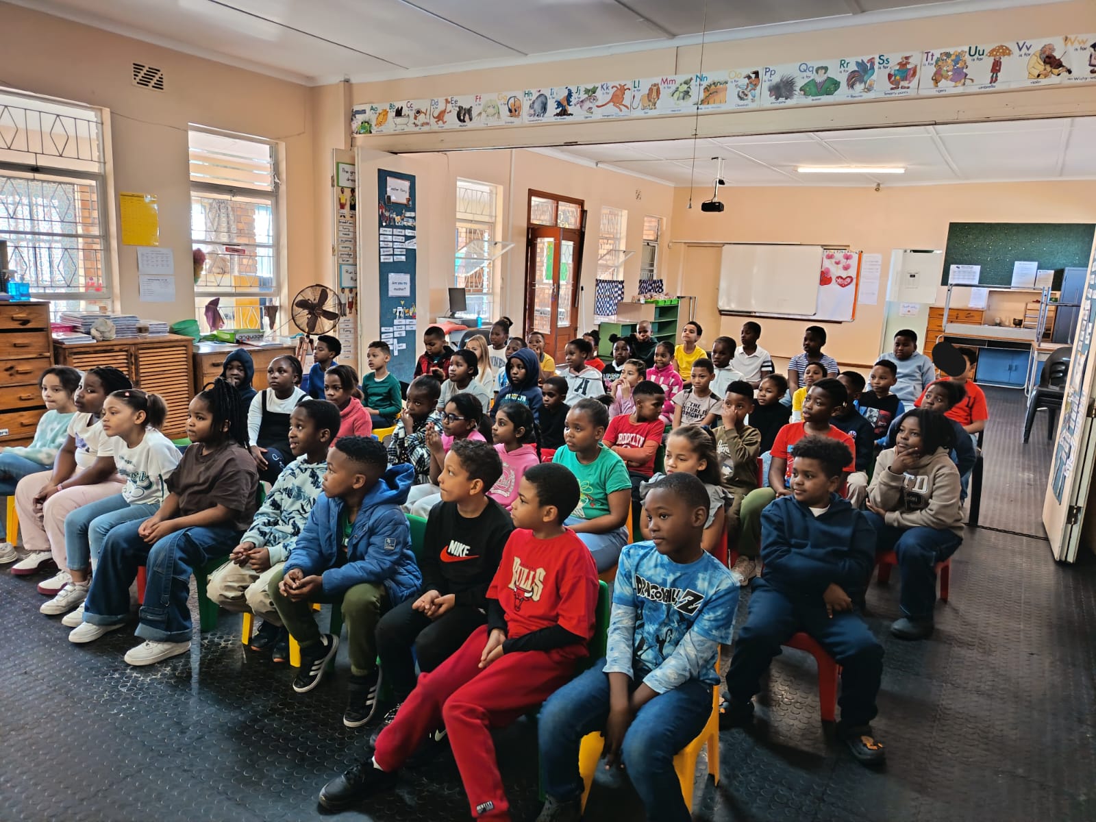 A group of young children sit on chairs in rows inside a classroom, facing forward and appearing attentive. The classroom has colorful decorations, posters, and large windows.