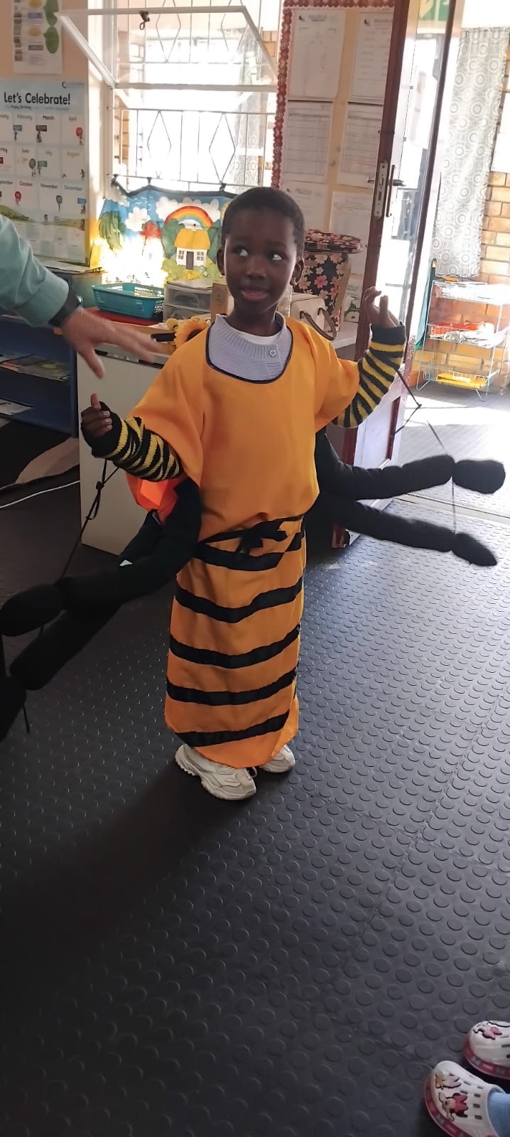 A child stands indoors wearing a yellow costume with black stripes and black fabric wings, resembling a bee. The child is looking up and holding their hands up. An adult's hand and part of another child are visible nearby.