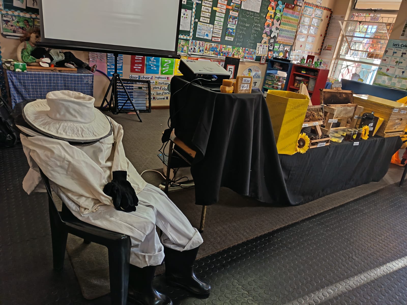 A classroom display features a mannequin in a beekeeper suit seated on a chair, with a table nearby holding beekeeping equipment, honeycombs, sunflowers, and educational posters on the walls.