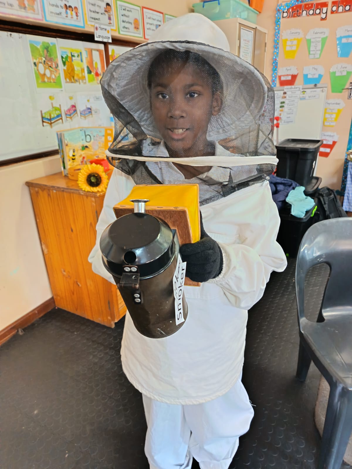 A child in a beekeeping suit holds a smoker and a rectangular yellow item in a classroom. Colorful posters and educational materials are visible on the walls and tables in the background.