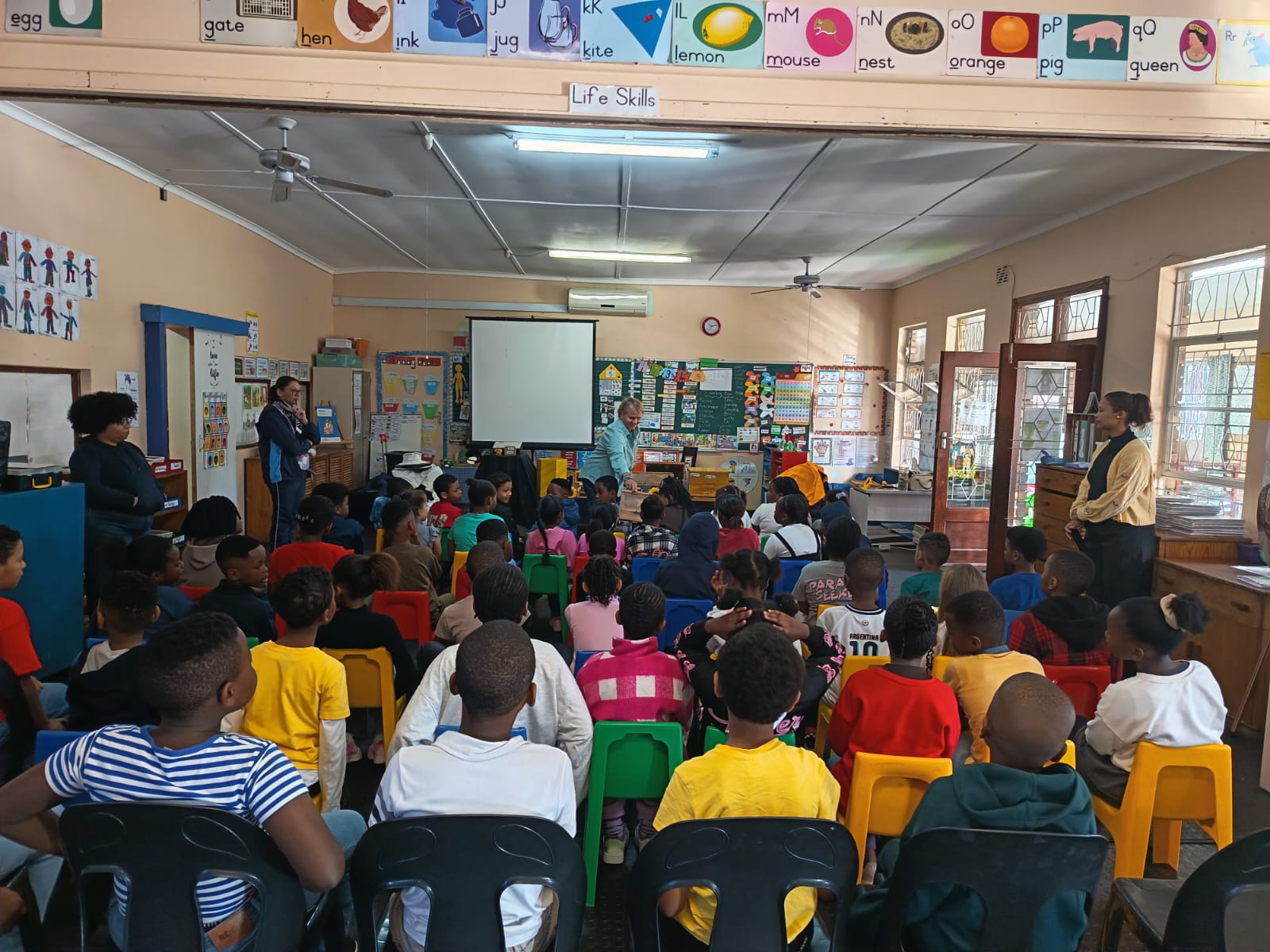 A classroom filled with young students sitting on chairs facing a teacher at the front, with several adults standing around. Educational posters and colorful decorations cover the walls. A projector screen is visible at the front.