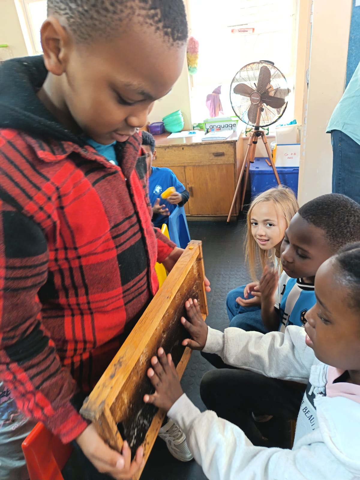 A boy in a red plaid jacket holds a wooden frame, while two seated children touch it and a girl with long blonde hair watches. The scene takes place in a classroom with a fan and various supplies in the background.