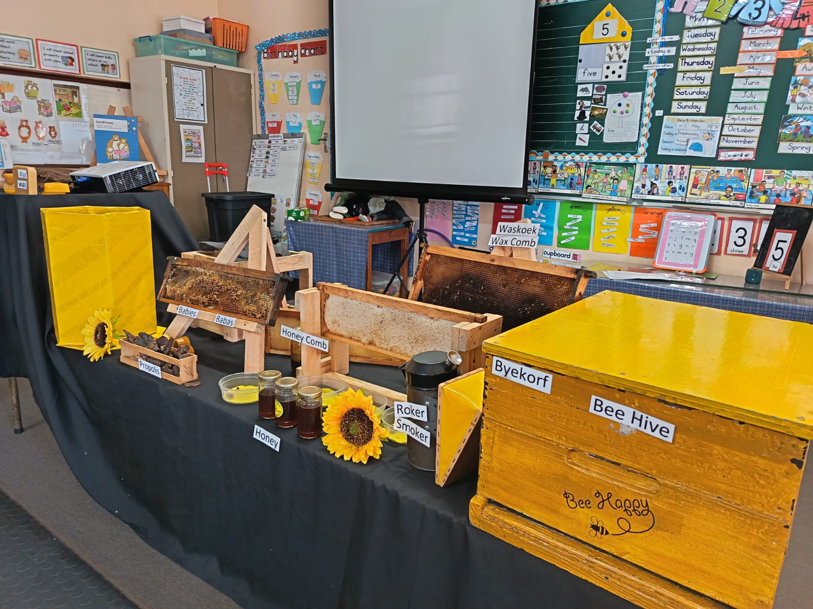 A classroom display features labeled beekeeping equipment, honey jars, honeycomb frames, a sunflower, and a yellow bee hive on a black table. Educational posters and a whiteboard are visible in the background.