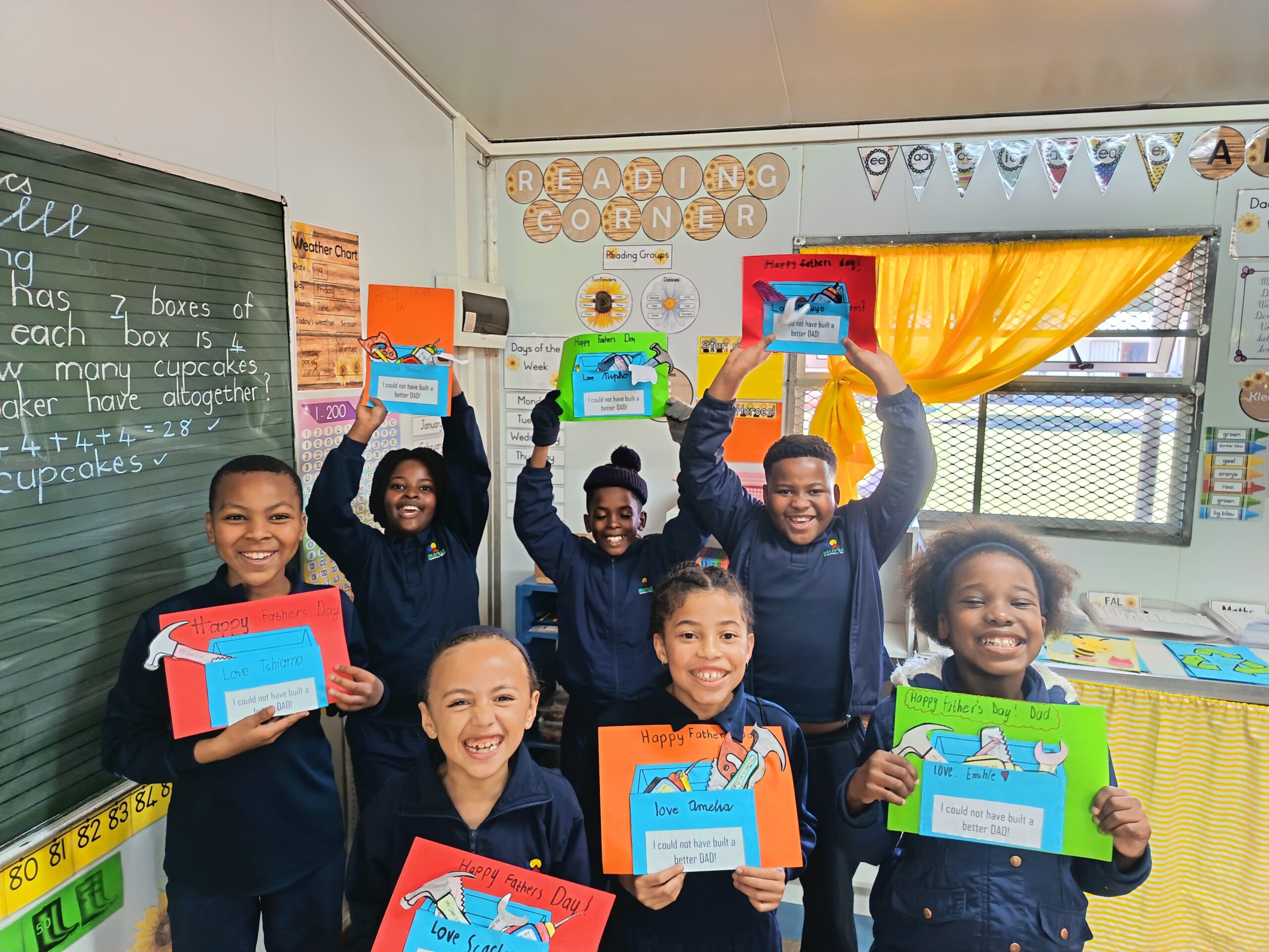 Seven children stand in a classroom, smiling and holding up colorful Mother’s Day cards they have made. Behind them are classroom decorations, posters, and a chalkboard with math written on it.