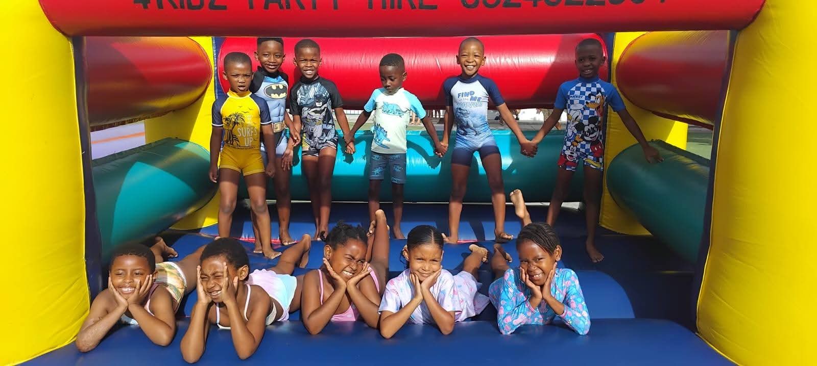 A group of children, some standing and some lying down, pose and smile on a colorful inflatable play structure outdoors, wearing casual clothes and swimwear.