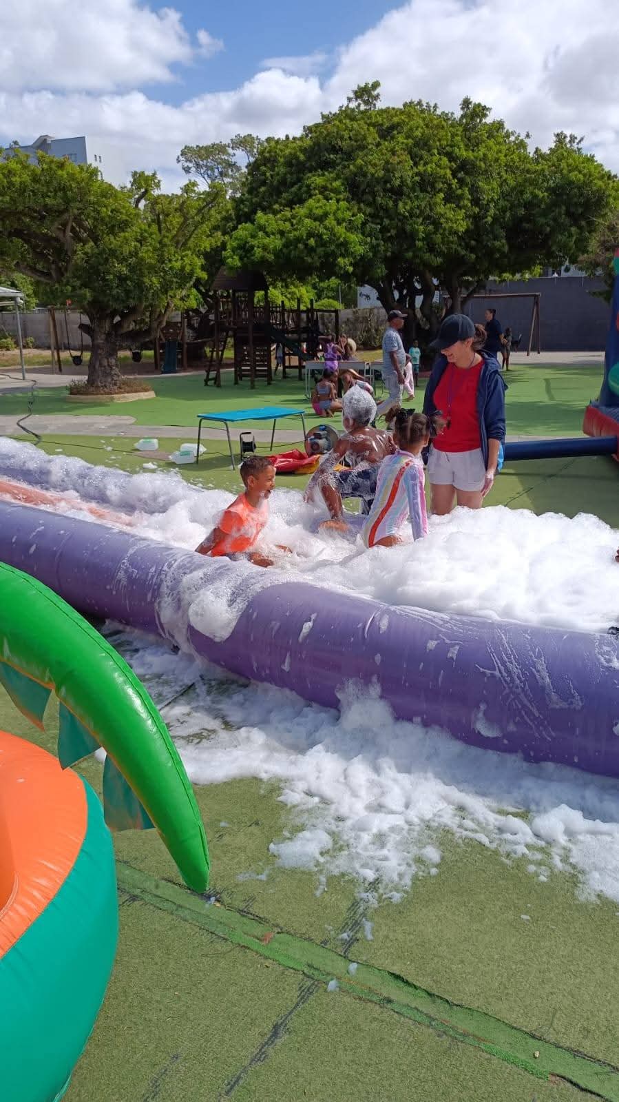 Several children play in foam on a purple inflatable structure outdoors, with adults supervising nearby. Playground equipment and trees are visible in the background on a sunny day.