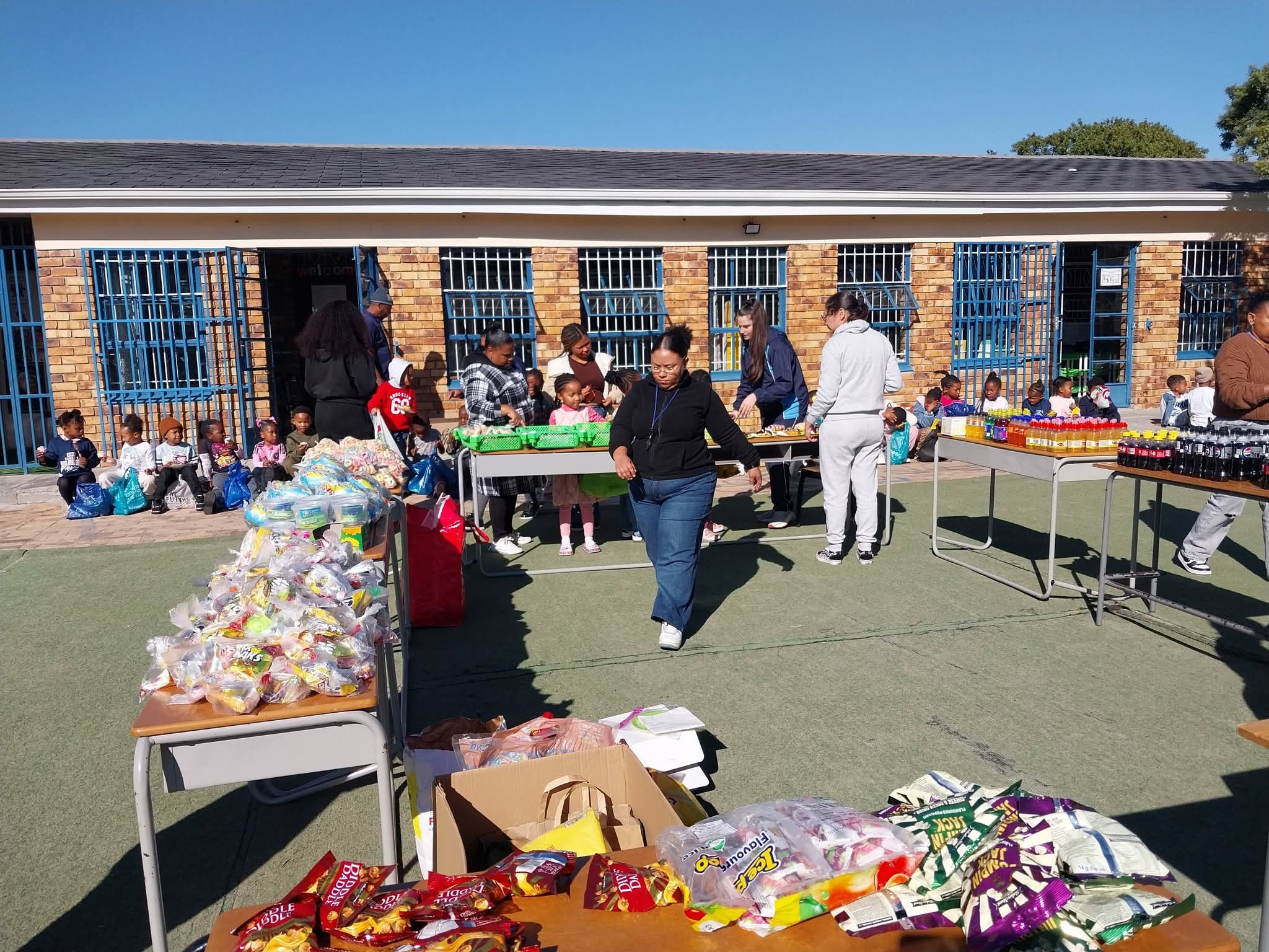 Tables with snacks and drinks are set up outdoors in front of a brick building with blue bars. Several people stand behind the tables, and a group of children sit on the ground in the background.