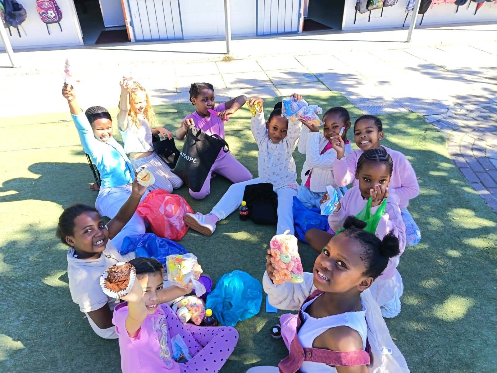 A group of children sit in a circle outdoors on a green mat, smiling and holding up colorful items or gifts. Various bags are around them. Some children are wearing light-colored clothes. The background shows buildings and shadows.