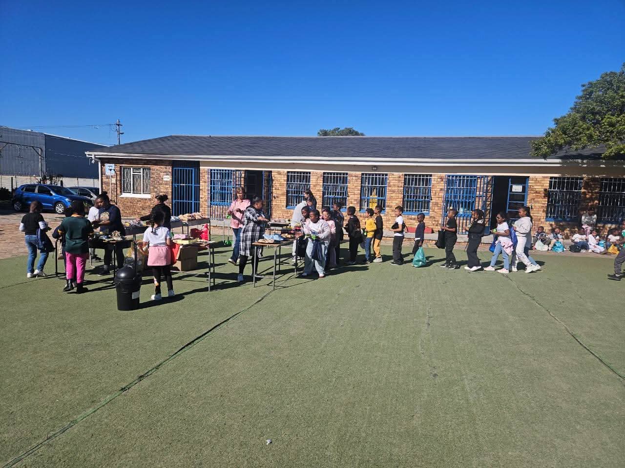 Children and adults stand in a line on a grassy field near a brick building with blue doors and windows. Tables with food or supplies are set up in the middle, and more children sit in the background. The sky is clear and blue.
