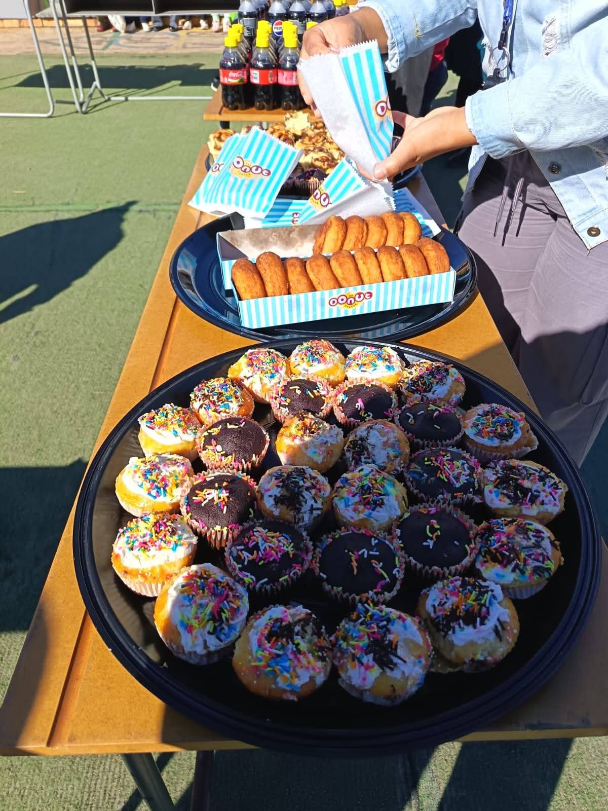 A table displays trays of mini cupcakes with sprinkles, a box of round pastries, and bottles of Coca-Cola. A person is serving pastries onto a plate. The setting appears to be outdoors.