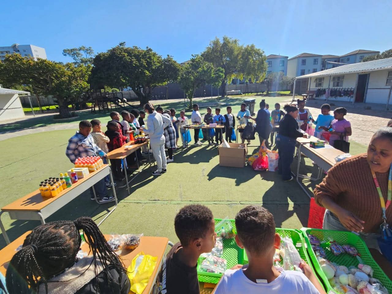 Children and adults stand in lines and behind tables with food and drinks on an outdoor sports court. Buildings and trees are visible in the background under a clear sky.