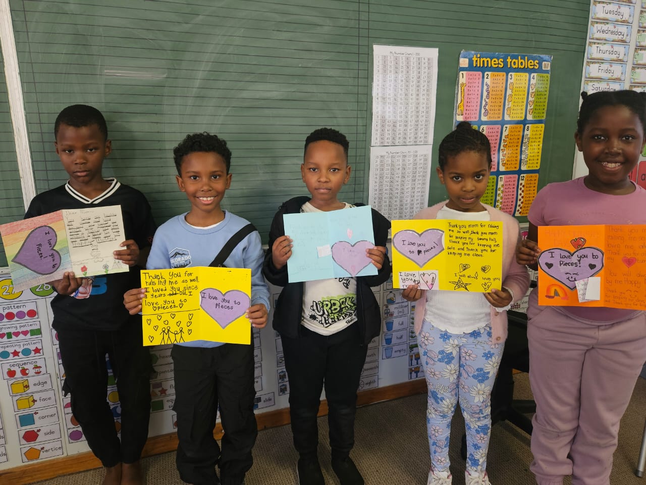 Five children stand indoors in front of a chalkboard and posters, each holding up colorful handmade cards with drawings and messages. The children are smiling and facing the camera.
