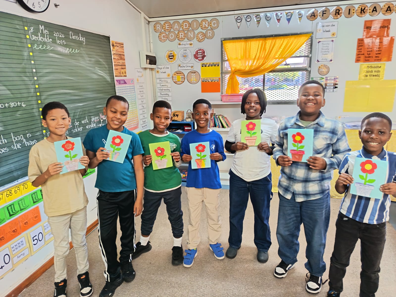 Six children stand in a classroom holding handmade cards with red flowers on them. They are smiling and posed in a row. The classroom has a green chalkboard, posters, and yellow-orange curtains.