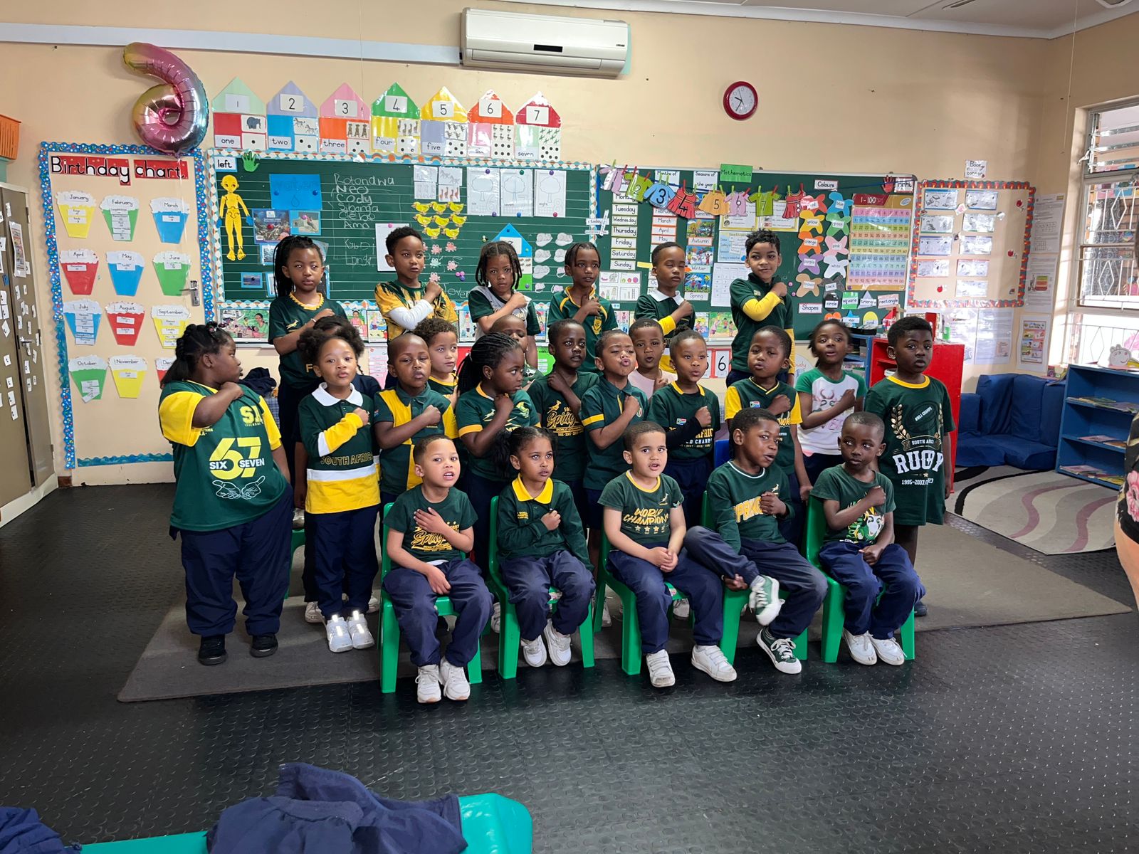 A group of young children in green and yellow sports uniforms pose for a class photo inside a colorful classroom, with some seated in front and others standing behind them. Classroom decorations and educational posters cover the walls.